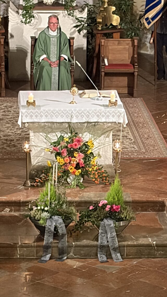 Pater Thomas Wagner im Hintergrund und vorne die Gestecke die am Kriegerdenkmal abgestellt werden.